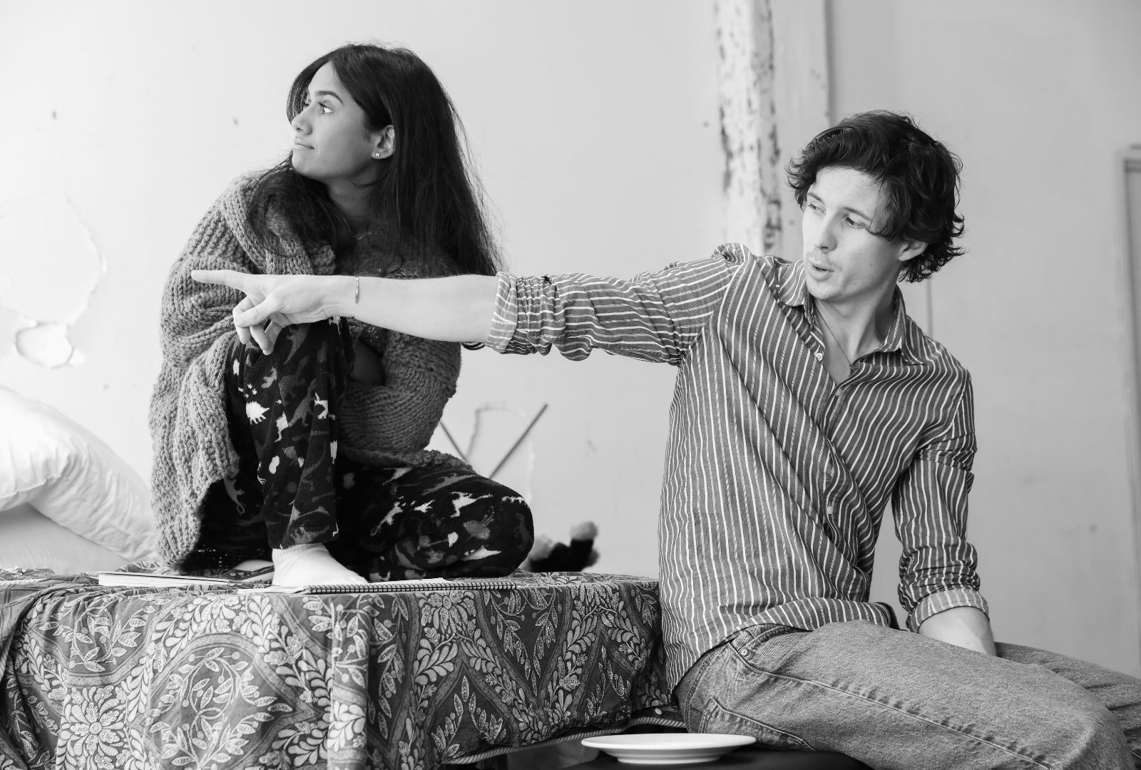 Black and white photo of Alyssa Peters crouched on a bed with Josh Hammond sitting below, pointing past her to something off screen.