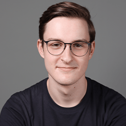 A young man with short brown hair and glasses, wearing a dark T-shirt, smiles softly while facing the camera against a plain grey background.