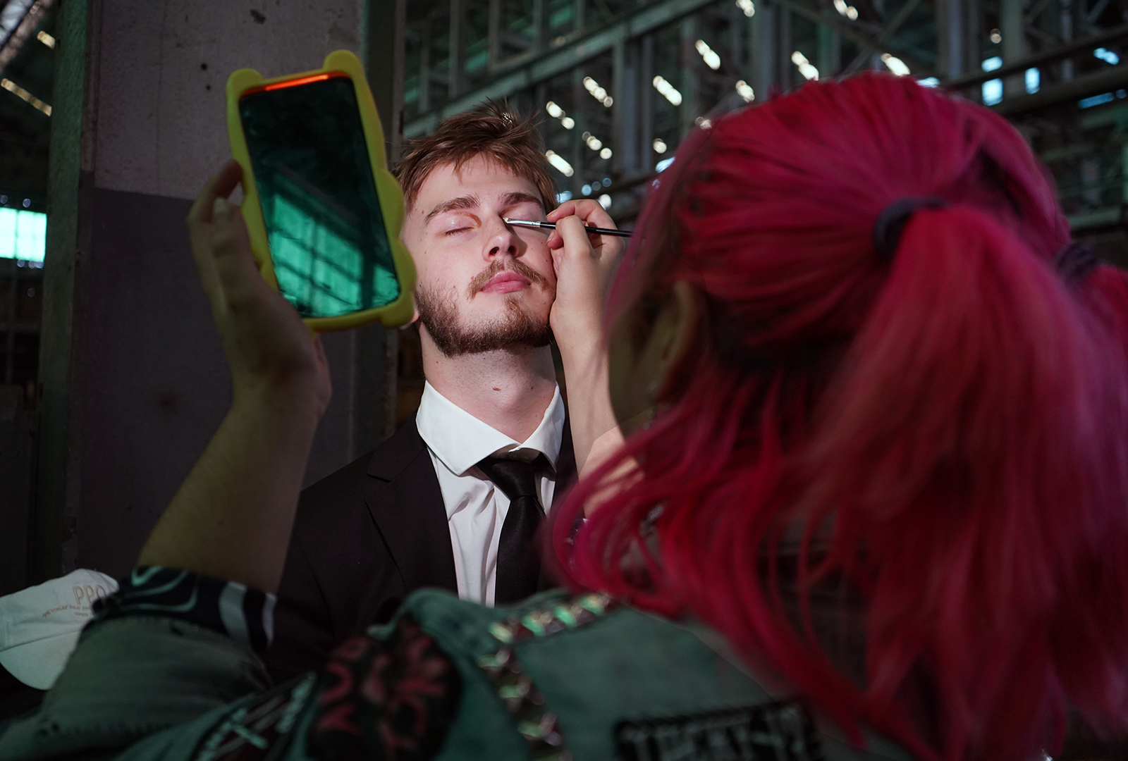 A person with pink hair applies eye make-up to a man in a suit, who has his eyes closed, while holding a yellow-framed mirror. They are in an industrial or warehouse-like setting with metal beams overhead.