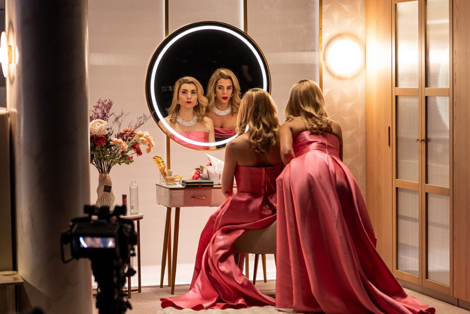 Two women are in soft, yellow light wearing pink gowns and large necklaces. They both look into a mirror with their backs to the camera.