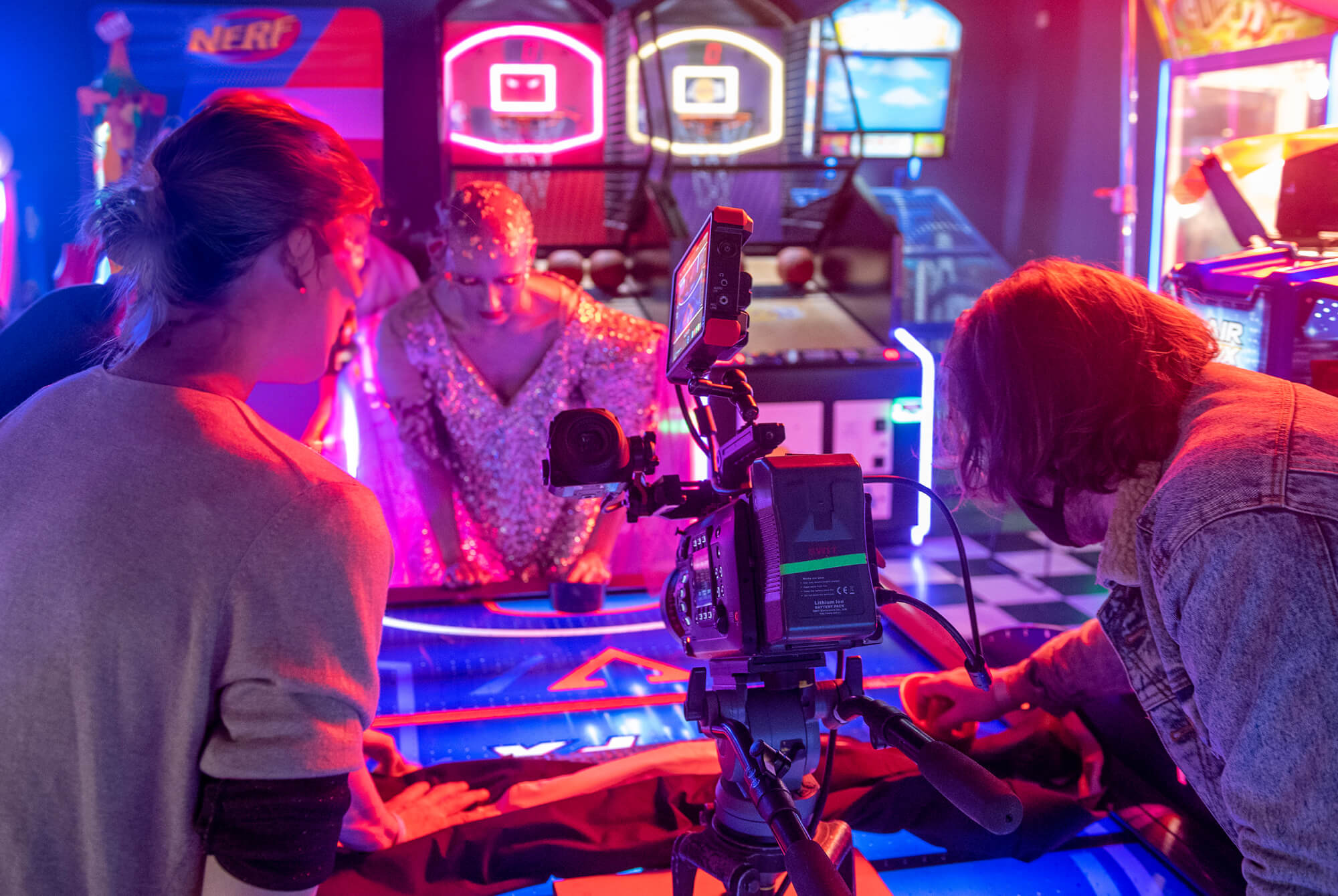 Three people are gathered around an air hockey table in an amusement arcade, with a camera and equipment set up in front of them. One person wears a sparkly outfit, while arcade machines and colourful lights fill the background.