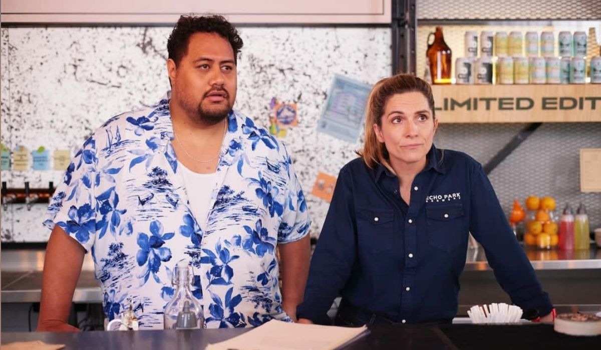 Two people stand behind a bar counter. The person on the left wears a blue floral shirt. The person on the right wears a dark blue shirt. They look attentive.