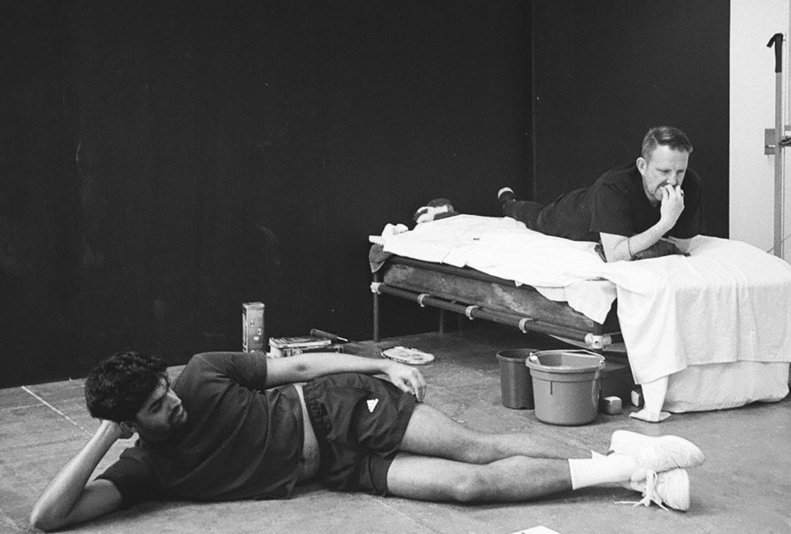 Black and white photo of two men, one laying on a bed the other on the ground.