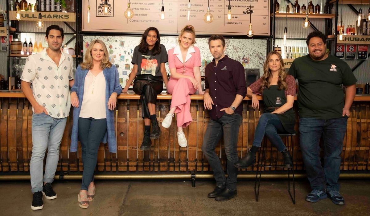 A group of seven people casually pose in front of a bar counter. They are smiling, creating a warm and friendly atmosphere. Bottles and a menu are visible behind them.