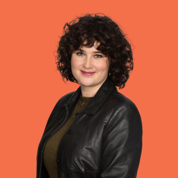 A woman with short, curly brown hair smiles at the camera. She is wearing a dark leather jacket over a brown top, standing against a solid orange background.