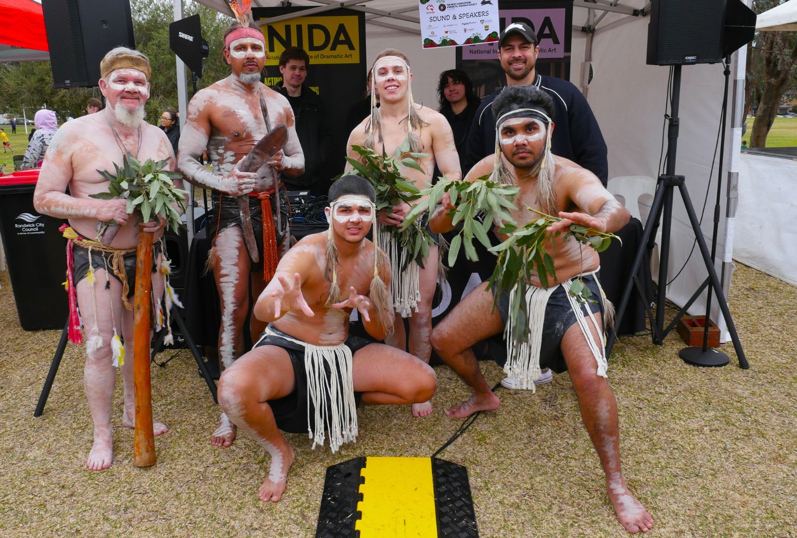 First Nations dancers in traditional paint and clothing, holding native leaves and instruments in front of the NIDA tent.