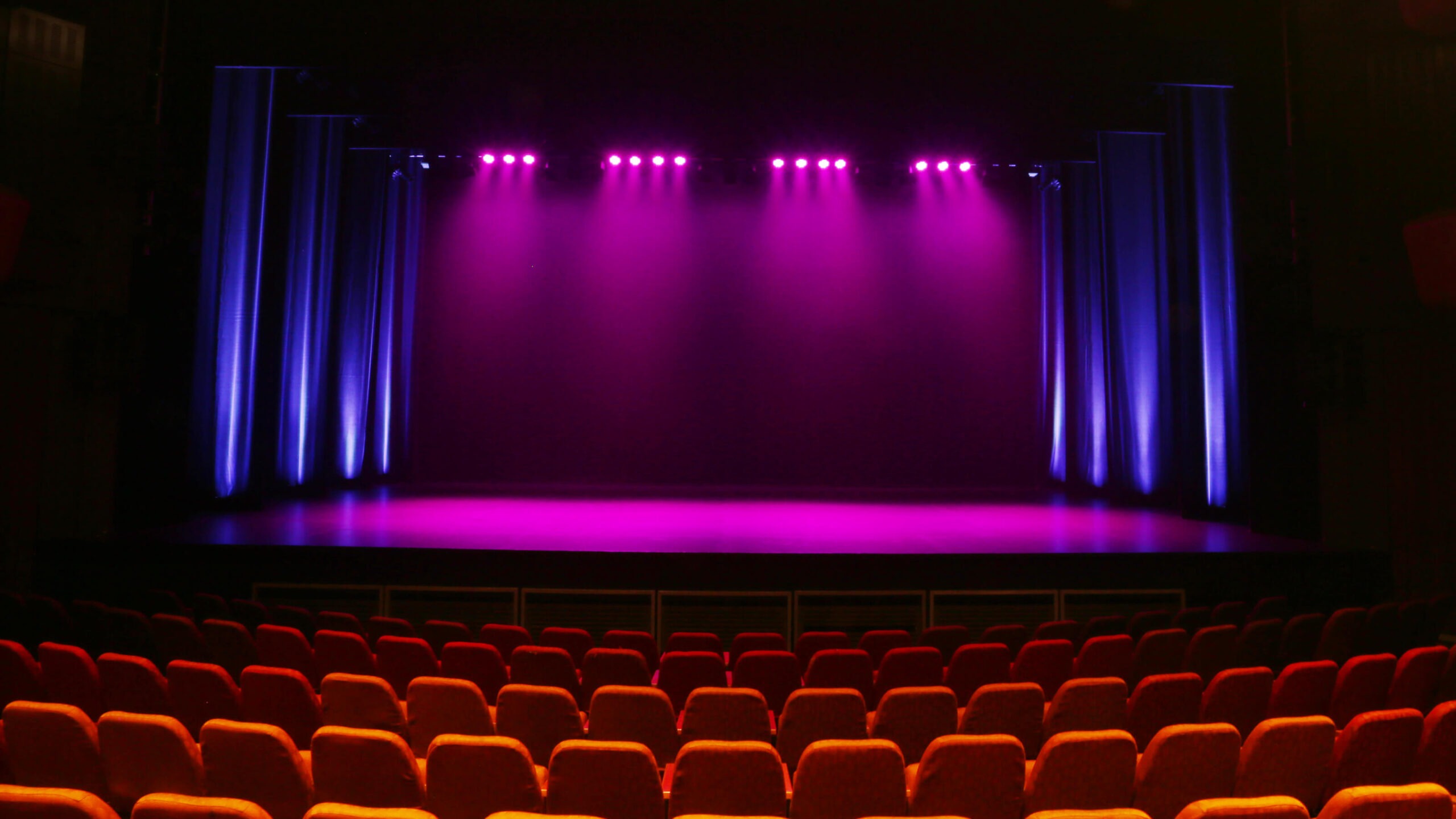 An empty theatre with rows of red seats facing a stage illuminated by purple and blue spotlights, with closed curtains and no performers present.