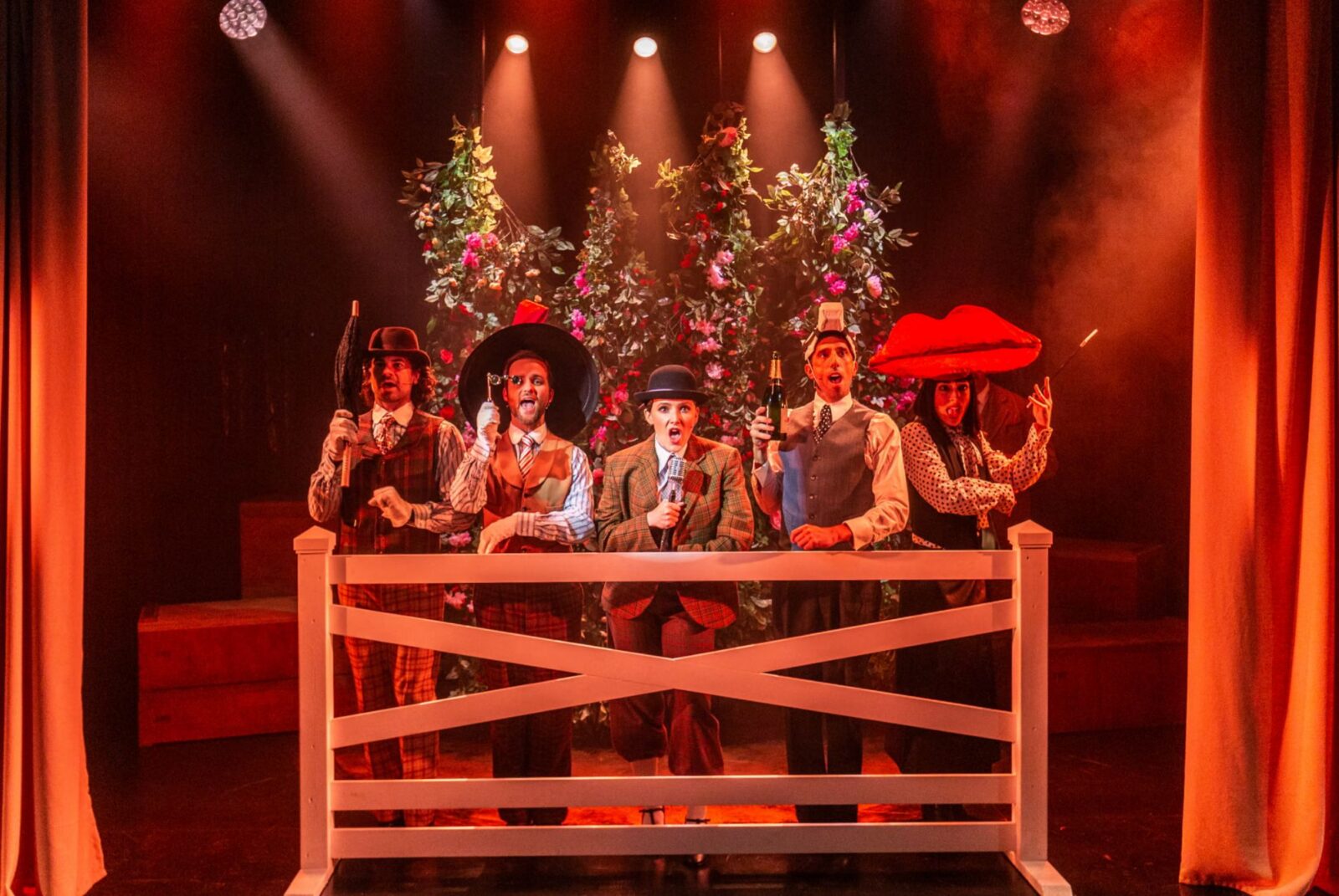 Five actors under red lighting in costumes standing behind a wooden gate.