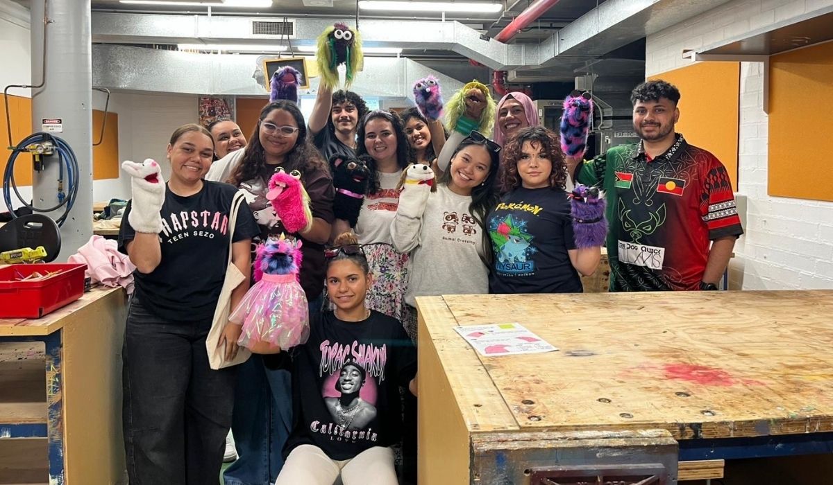 A group of smiling people holding colourful hand puppets stands in a workshop. The mood is cheerful and creative, with puppet-making materials around.
