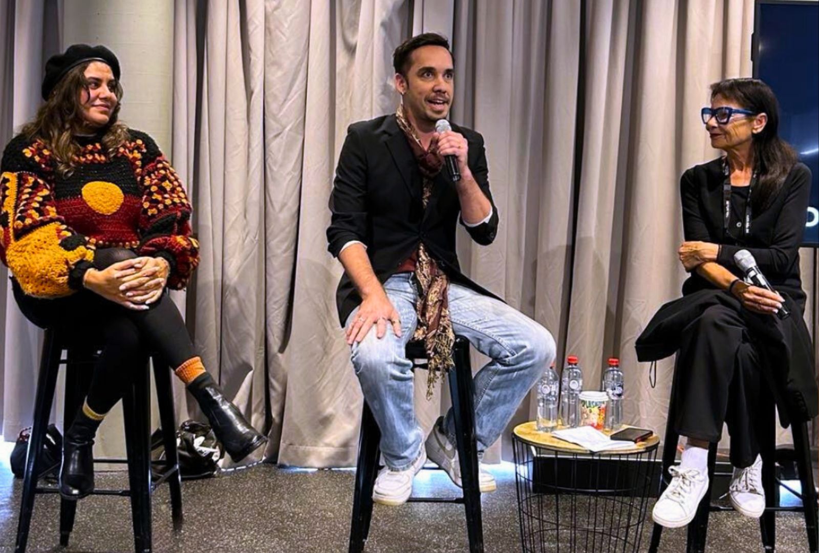 Three people with microphones sitting in front of a crowd talking.