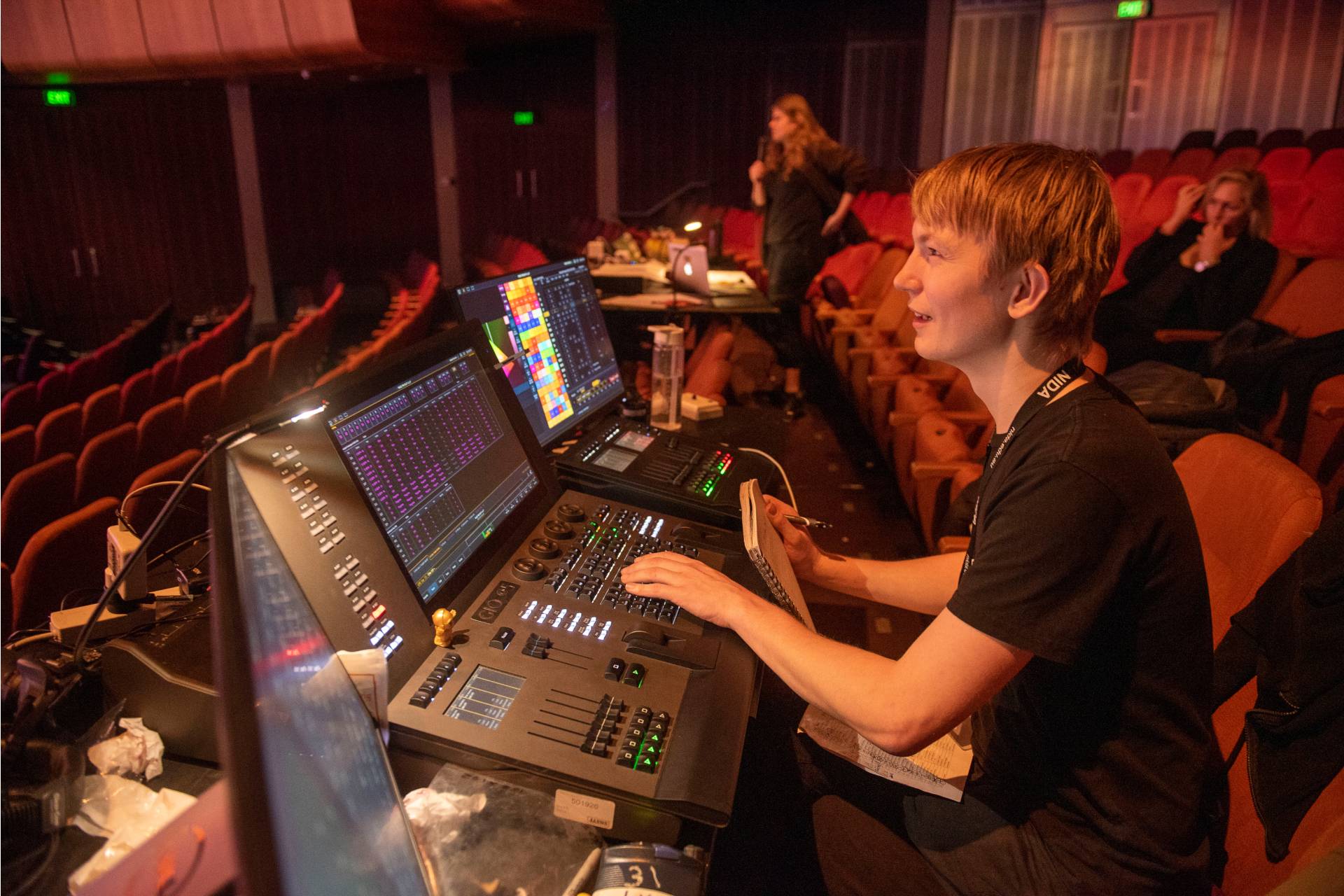 A person operates lighting and sound control panels in a theatre, focusing on screens and controls. Other people are seated nearby in the dimly lit auditorium.
