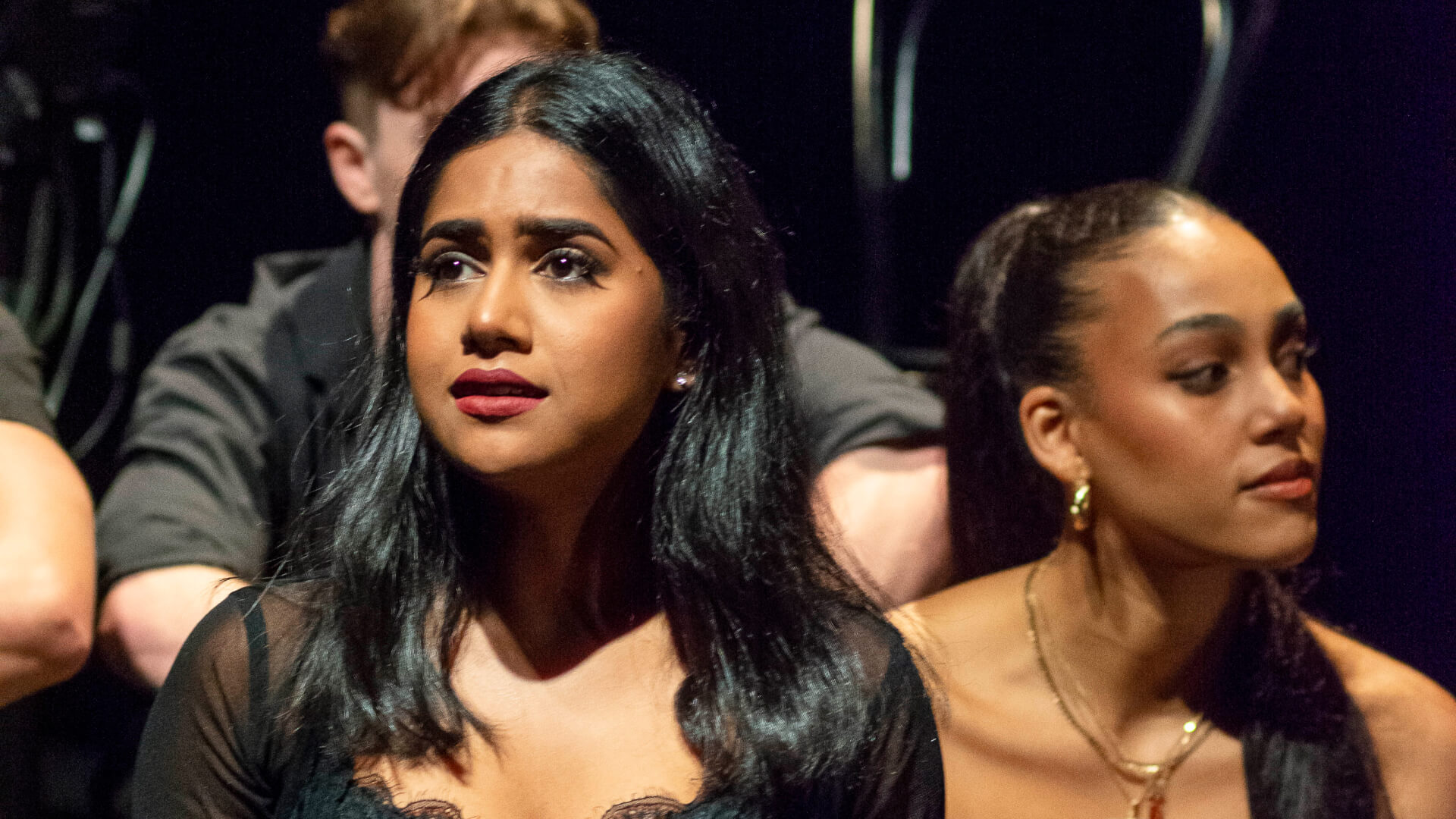 Two women sit side by side in what appears to be a theatre or performance setting. The woman on the left looks emotional, whilst the woman on the right looks off to the side with a neutral expression.