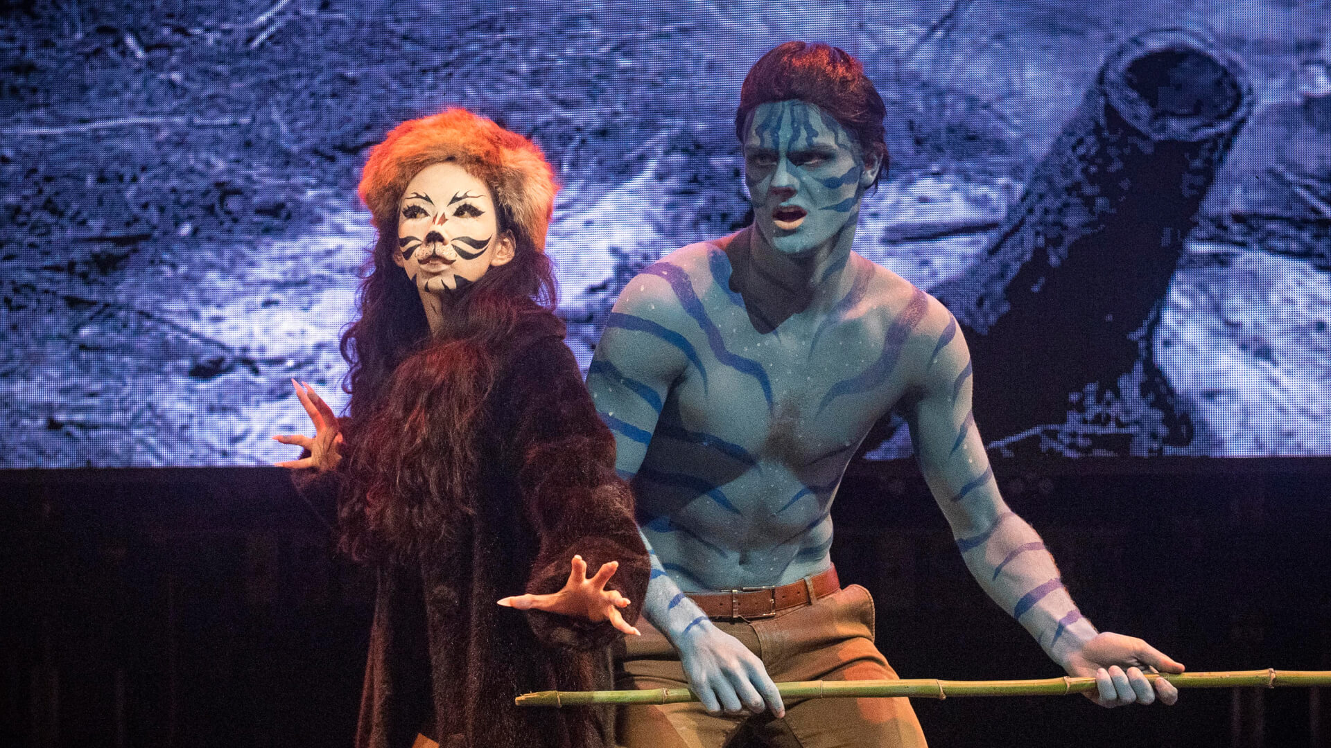Two actors perform on stage: one dressed as a cat with face paint and fur, the other bare-chested with blue body paint and stripes, holding a staff. A dark, dramatic backdrop is visible behind them.